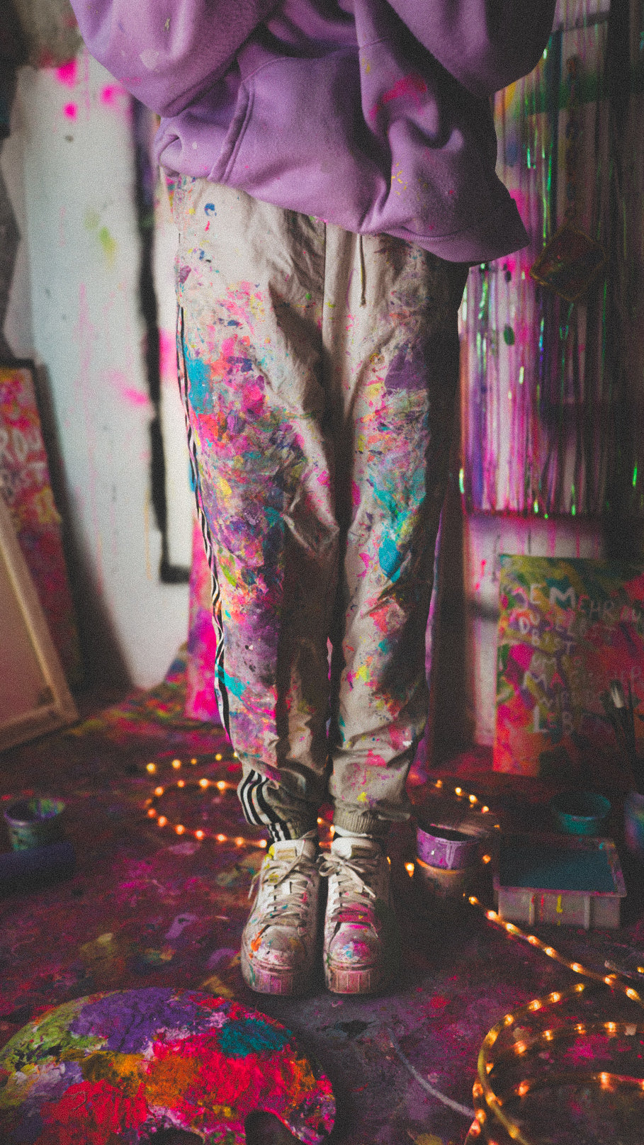 Künstlerin im Atelier mit bunter, farbbespritzter Kleidung und Sneakers. Kreatives Chaos aus Neonfarben, Leinwänden und Lichterketten. Kunst entsteht mit Leidenschaft und Farbe bei Natalie Irber. Künstlerisches Schaffen und Inspiration im kreativen Arbeitsraum.